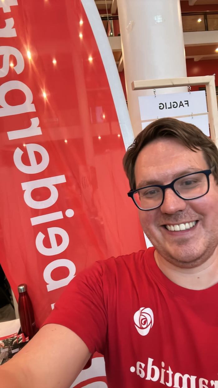 Karl Henrik står på stand og tar smilende en selfie med parti-t-skjorte på og beachflagg for Arbeiderpartiet i bakgrunnen på foreningsdagen i Georg Sverdrups hus, Blindern på Universitet i Oslo
