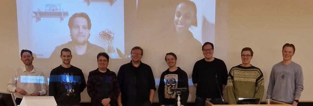 Deltakere på årsmøtet 2025 smiler til kamera med både fysisk og digital deltakelse i et auditorium på Blindern ved Universitetet i Oslo mot hvit bakgrunn.