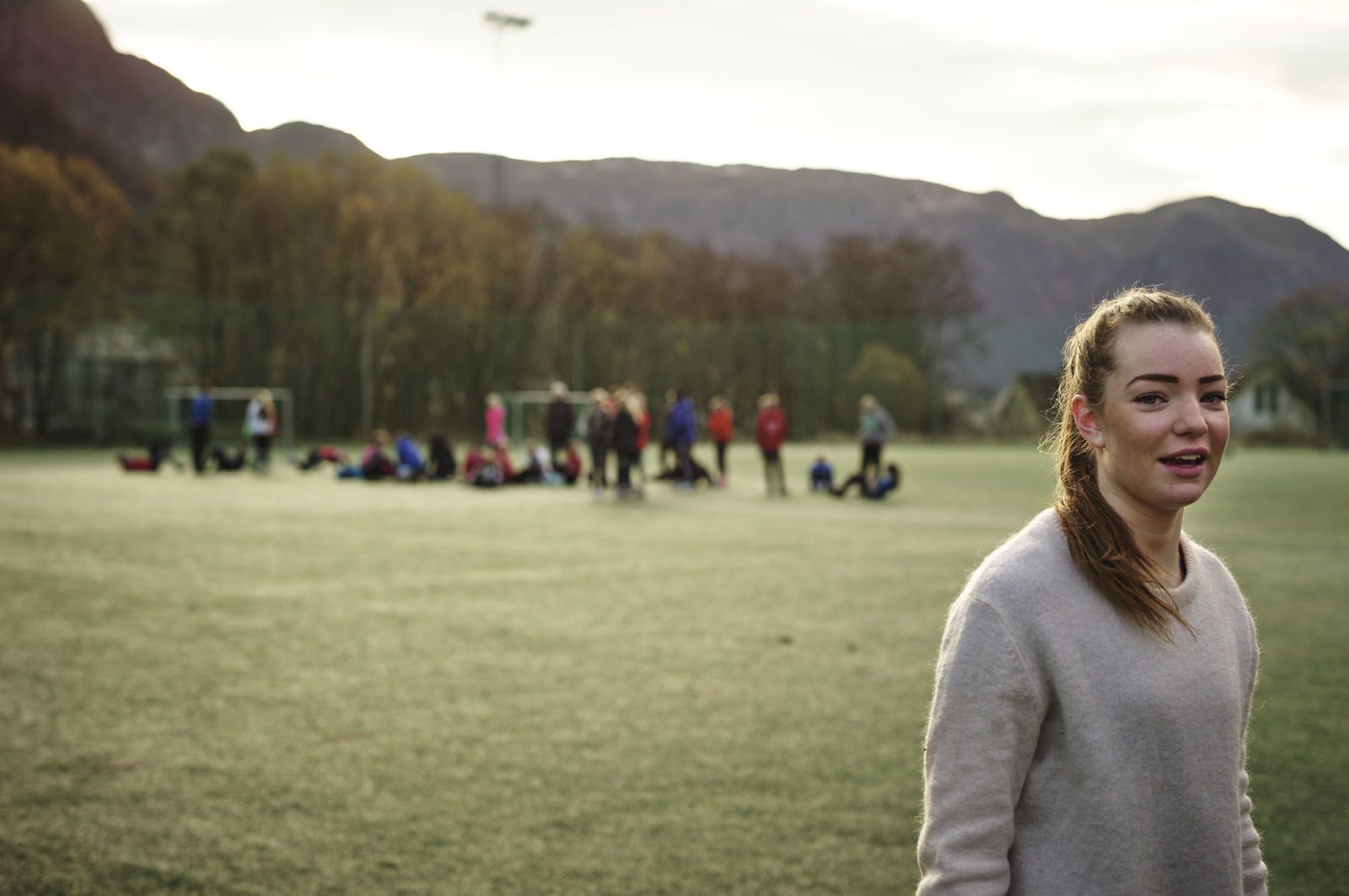 I forgrunnen ser vi en jente, i bakgrunnen står et fotballlag på banen. Foto: Øivind Haug
