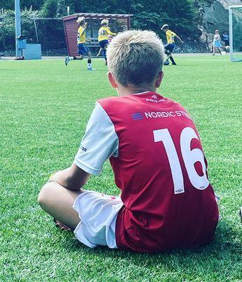 Gut som er fotballspelar sit på graset og ser på andre som spelar fotball
