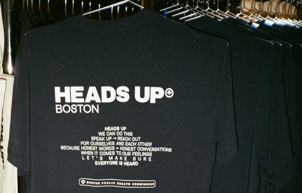 Navy blue t-shirt on a rack displaying "Heads Up Boston" and a message encouraging open conversations about feelings, issued by the Boston Public Health Commission.