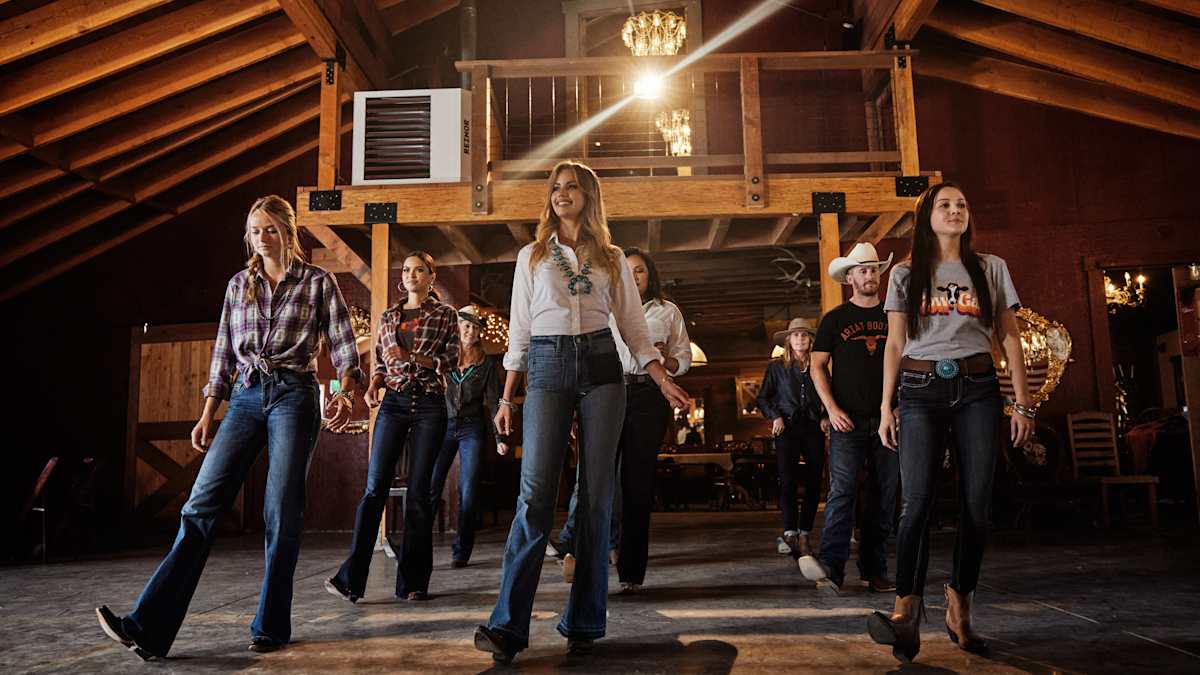 A group of men and women in Western attire line dancing in a barn with glowing chandeliers.
