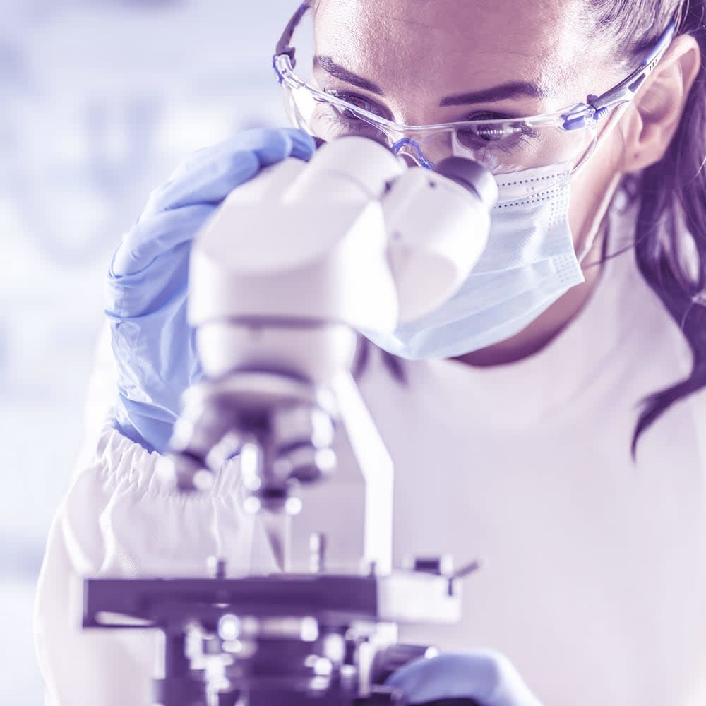 lab technician in protective glasses, gloves and face mask looking in a microscope in laboratory