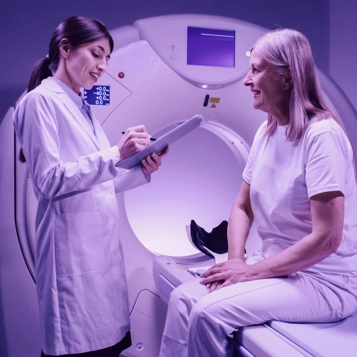 Female doctor is standing near patient and explaining procedure. Female patient is smiling and listening to doctor carefully. Medical worker is holding tablet and talk to patient.