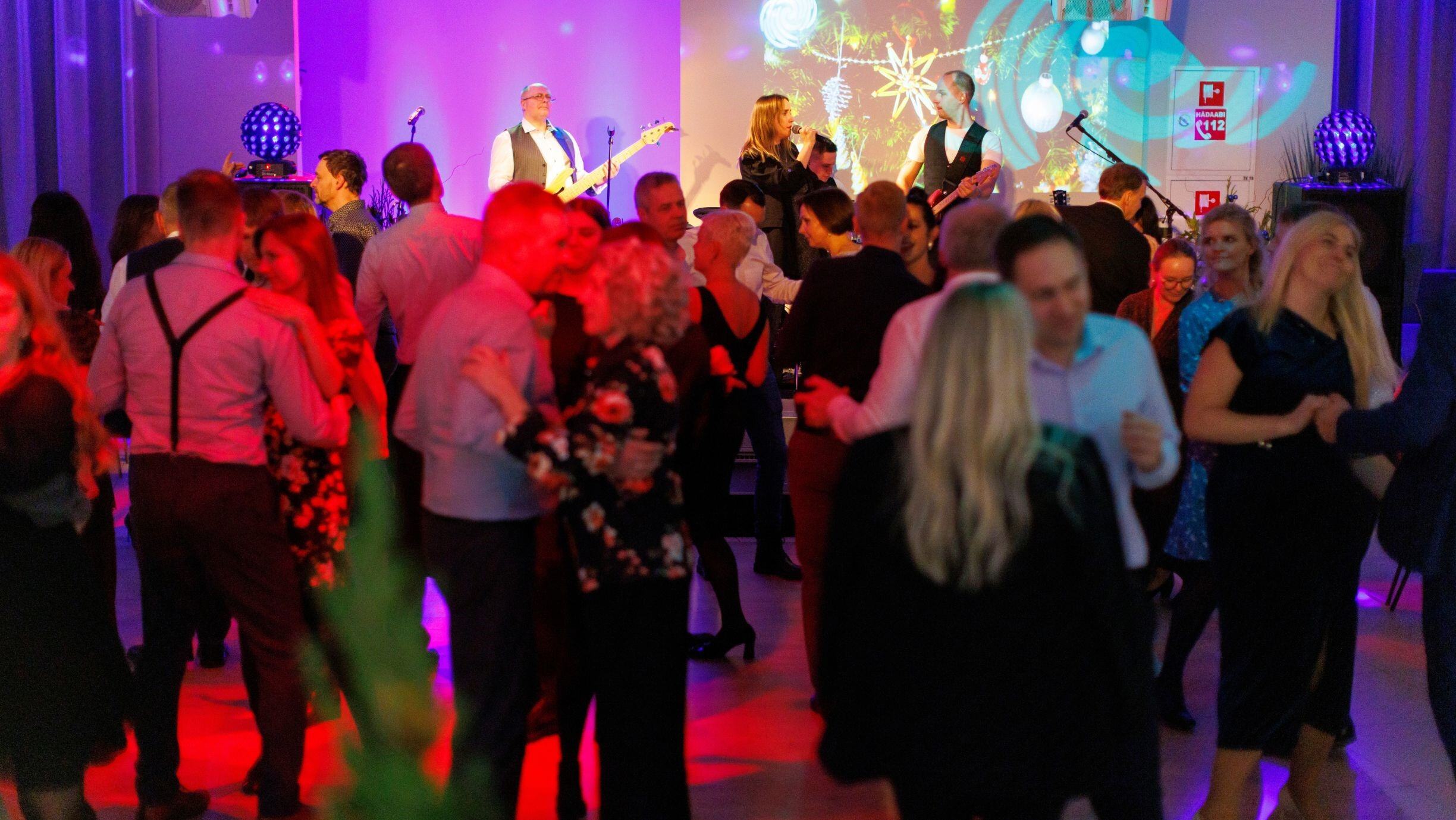 Guests are dancing at Nero restaurant. Most of them have their backs to the camera, a band is performing on stage, and the atmosphere is cheerful, with bright red and yellow tones.