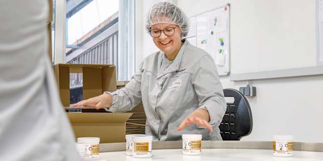 Mitarbeiterin in Hygienekleidung verpackt Dosen „Kräuterhof® Pferdebalsam“ an einem runden Arbeitstisch.