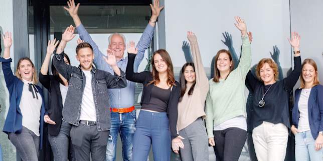 Gruppenporträt von neun Personen vor einem Gebäude, mehrere winken