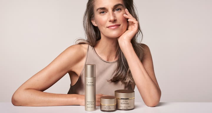 Femme au maquillage naturel appuyée sur une table à côté de soins M. Asam, flacon de sérum et deux pots de crème dans un décor neutre de studio
