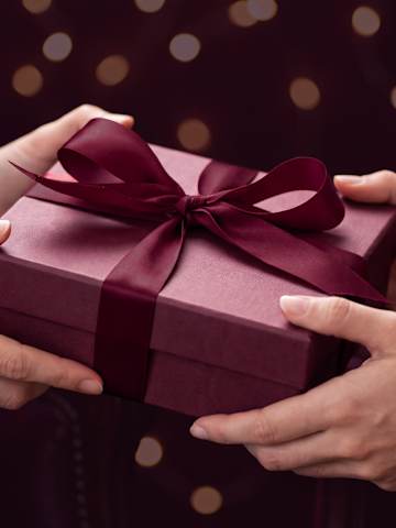 Geschenkübergabe einer bordeauxroten Geschenkbox mit Satinschleife vor dunklem Hintergrund
