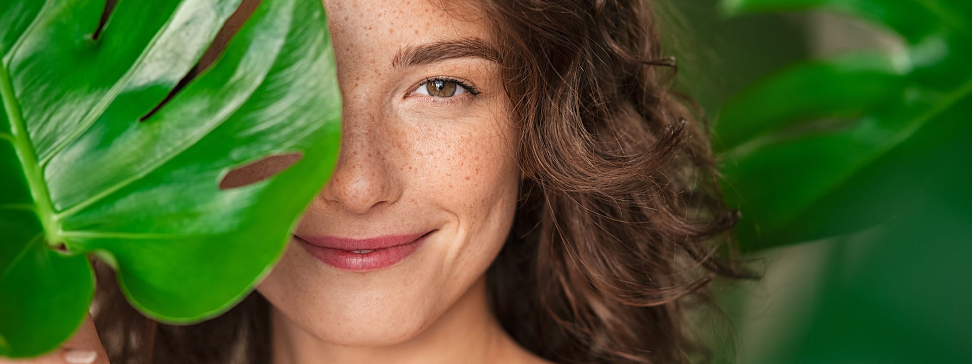 Frau mit lockigem Haar und Sommersprossen hält ein großes Monstera-Blatt vor ihr Gesicht, ein Auge ist sichtbar.