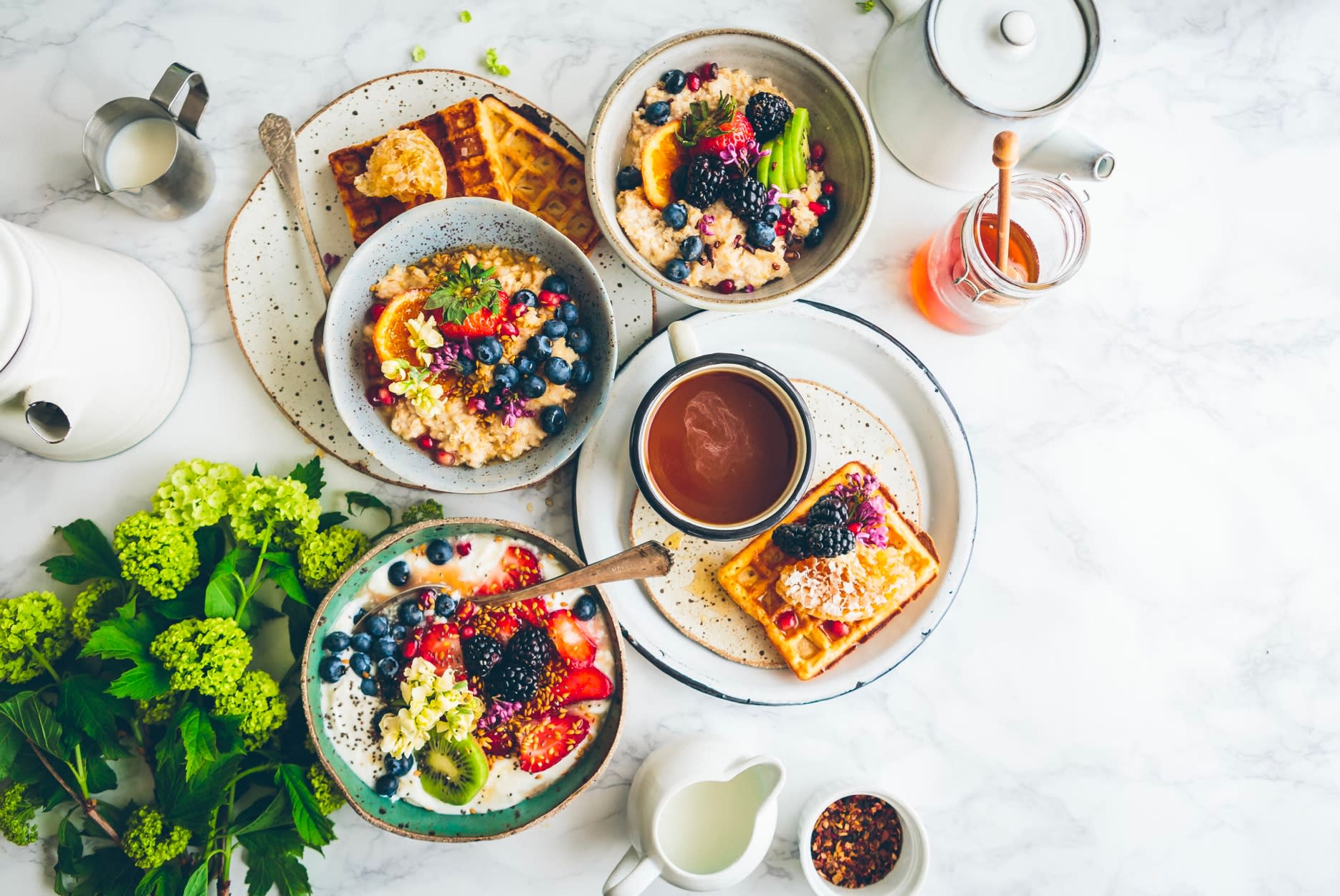 A variety of colorful and appetizing breakfast dishes, including waffles, oatmeal, and fresh fruits, are arranged on a white surface, accompanied by various condiments and utensils.