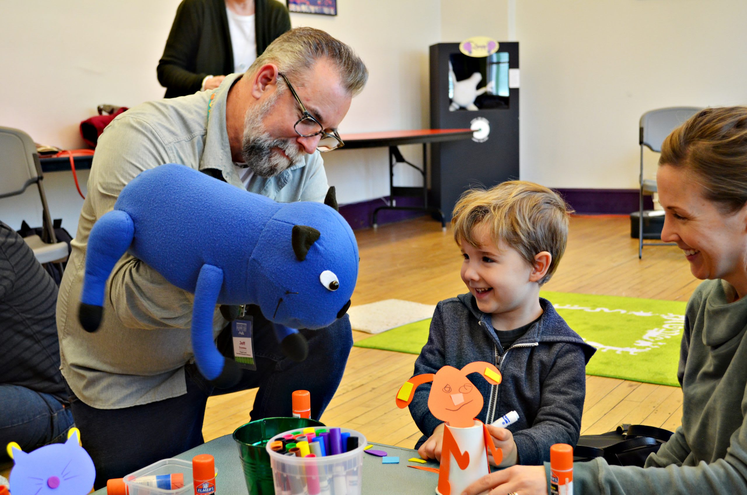 A puppeteer from the Center of Puppetry Arts, in Atlanta Georgia, shows a blue cat puppet to a smiling little boy and his caregiver during a workshop.