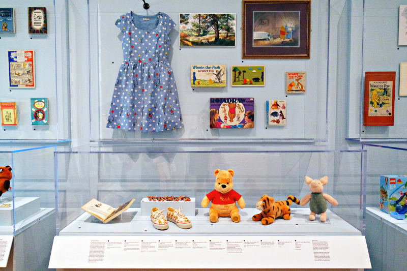 This display from the Winnie-the-Pooh: Exploring a Classic exhibit at the High Museum of Art Atlanta features Winnie-the-Pooh, Tigger, and Piglet stuffed animals, a dress, shoes, artwork, games, books, and more.