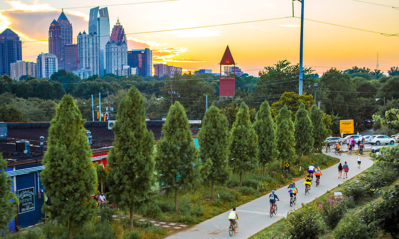 Cyclists ride along a tree-lined path at sunset with a city skyline in the background in Atlanta, Georgia.