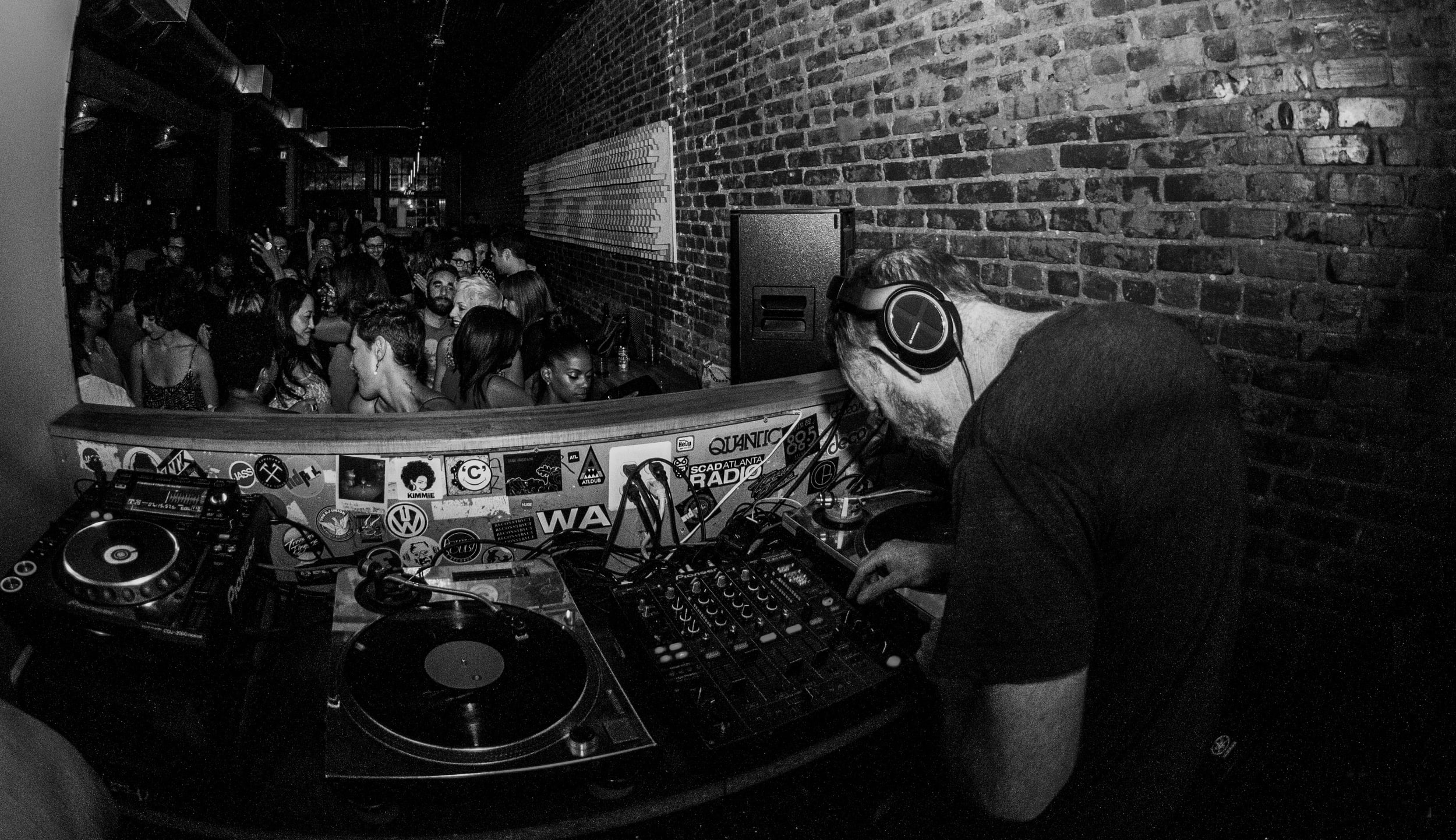 A DJ wearing headphones mixes music in a dimly lit club with a brick wall and dancing crowd in Atlanta, Georgia.