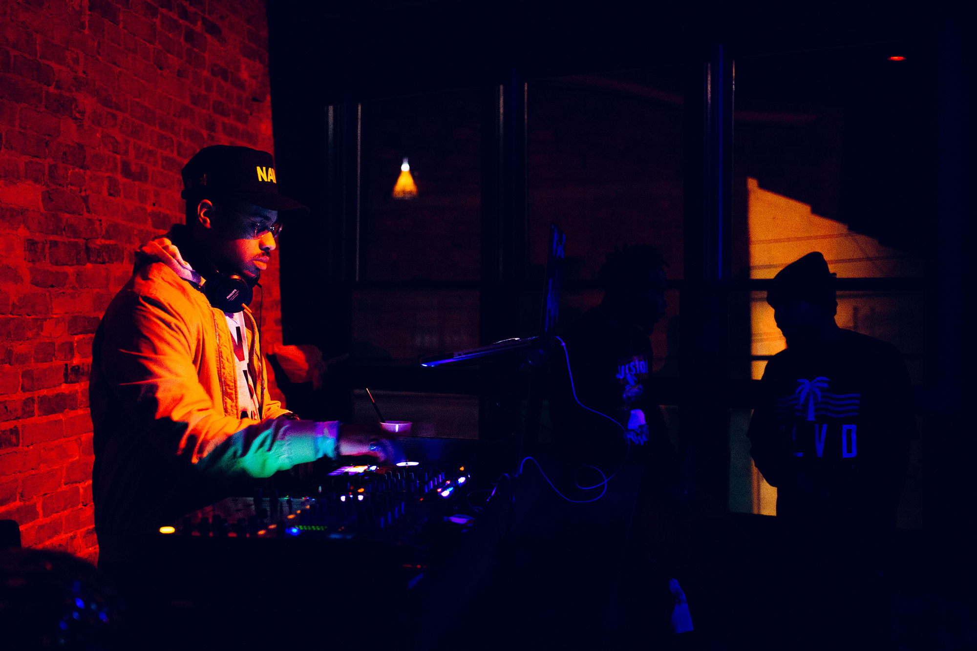 A DJ wearing a yellow jacket and a cap with 'NA' on it is performing in a dimly lit room with a brick wall in Atlanta, Georgia.