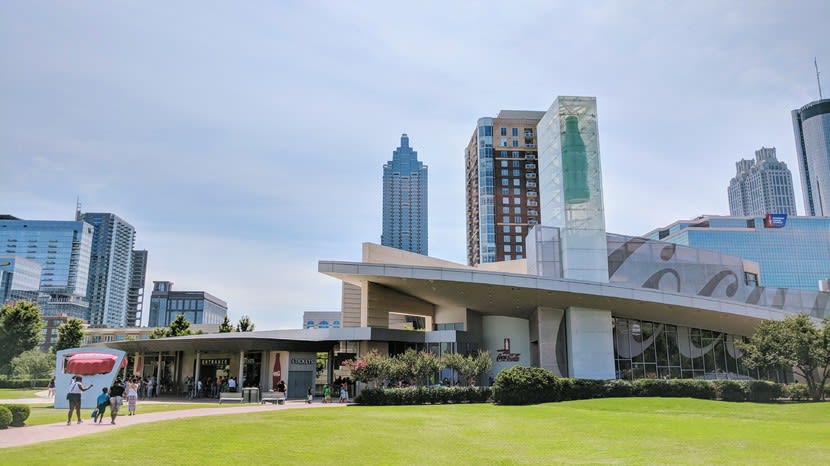 A modern building with a large Coca-Cola logo and a glass bottle sculpture stands in a grassy area with people walking around in Atlanta, Georgia.