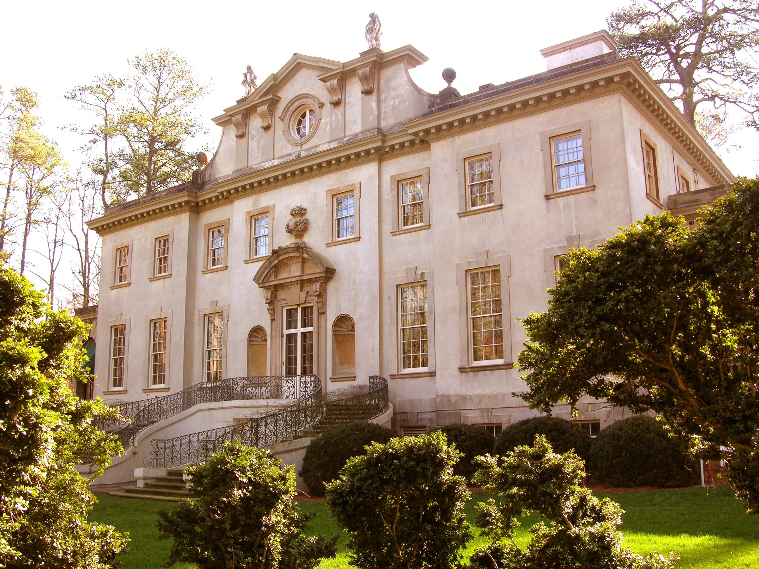 A grand, pale yellow mansion with a curved staircase and ornate details surrounded by lush greenery in Atlanta, Georgia.