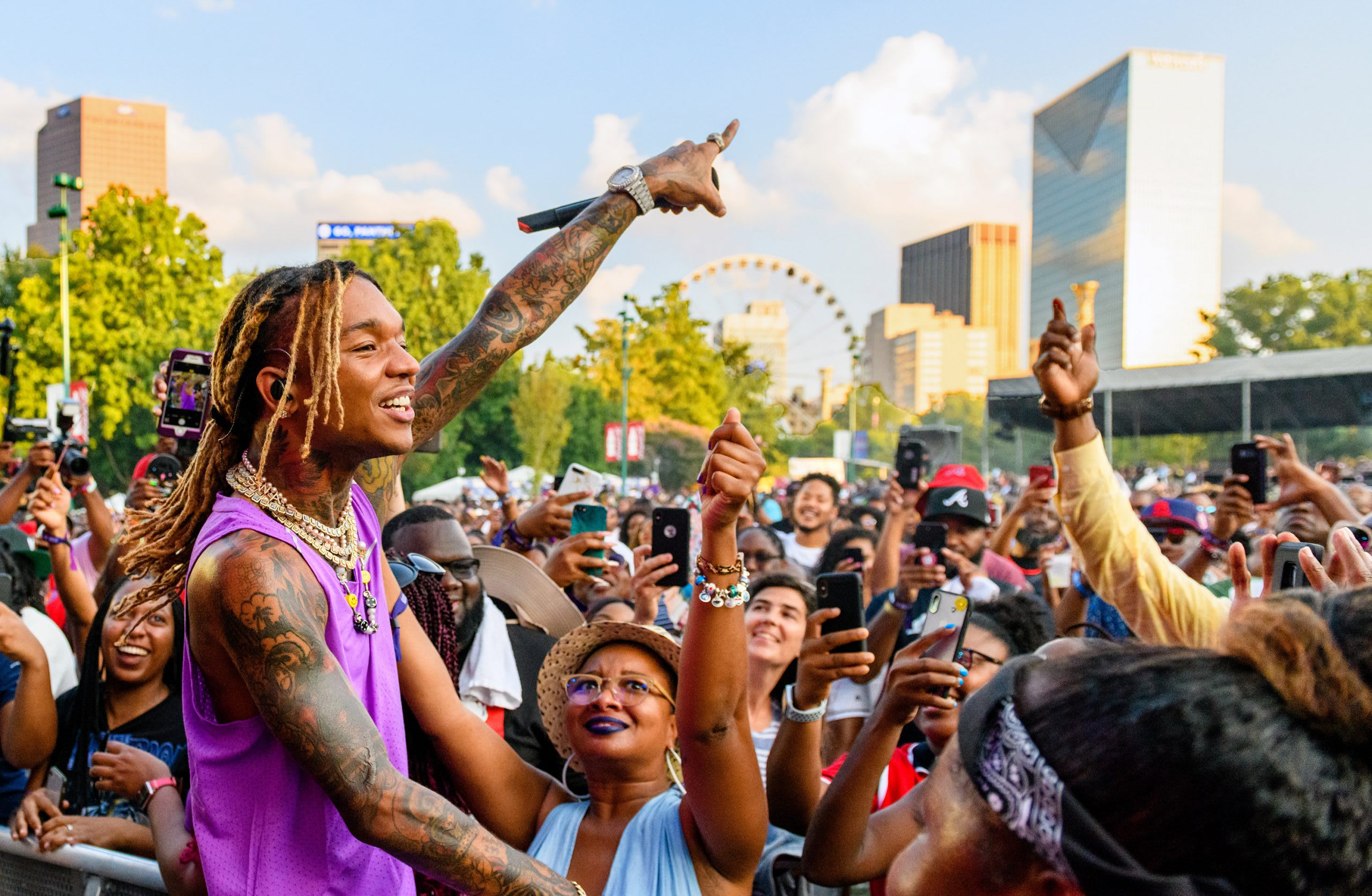 A large crowd at an outdoor concert, with a tattooed performer in purple raising his arm, surrounded by enthusiastic fans in Atlanta, Georgia.