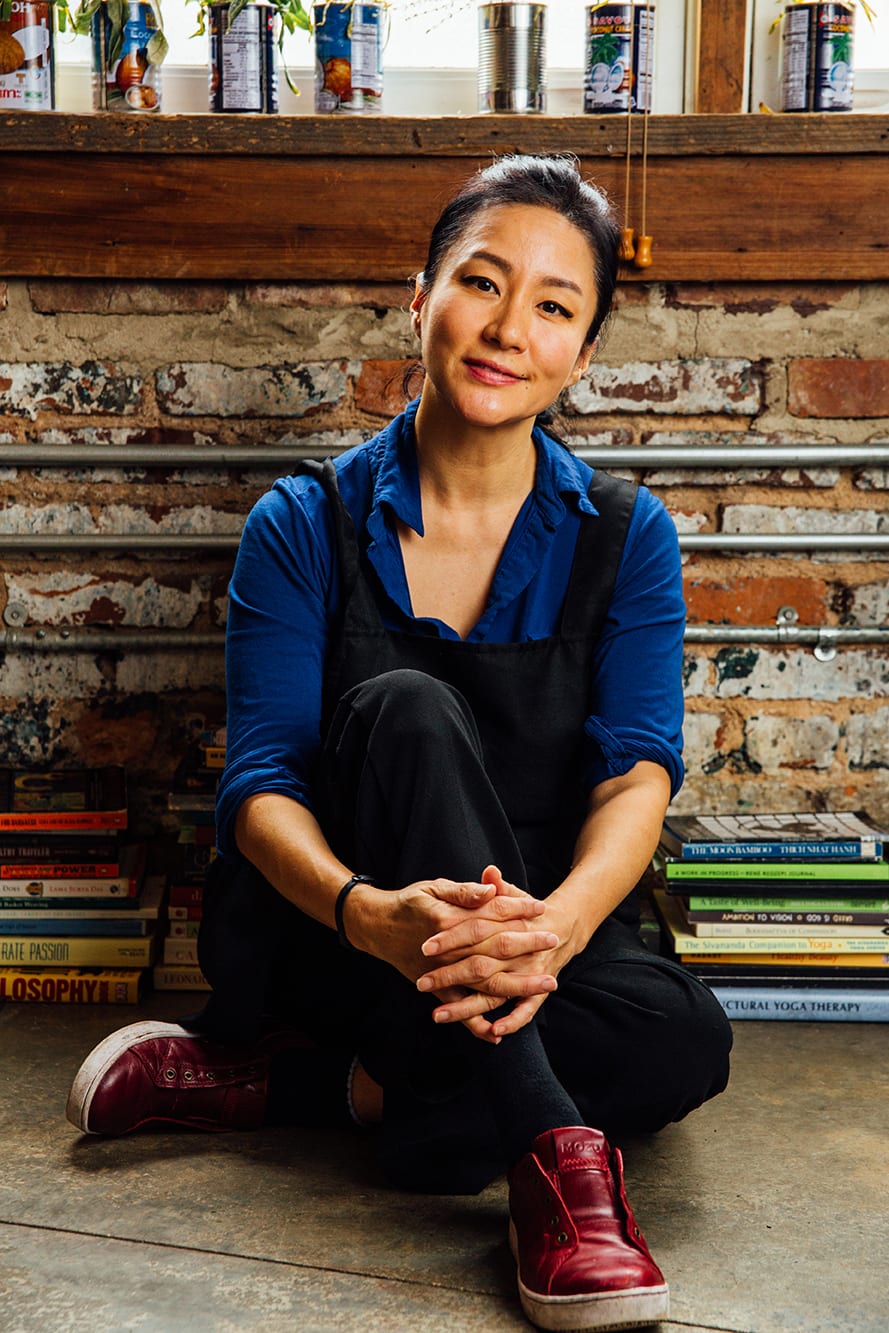 A woman sits cross-legged on a concrete floor, surrounded by books and cans on a shelf in Atlanta, Georgia.