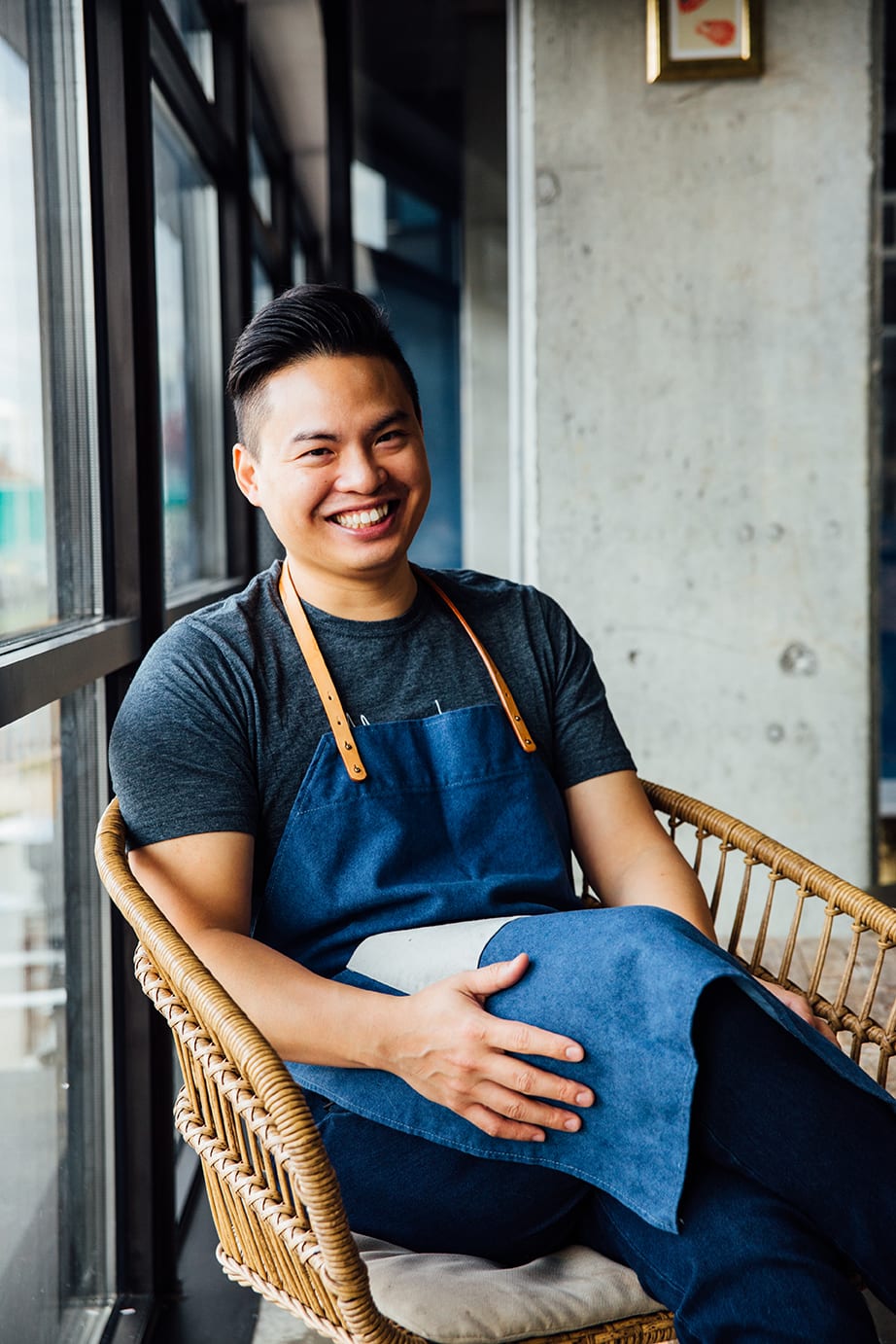A smiling man wearing a denim apron sits in a wicker chair by a window in Atlanta, Georgia.