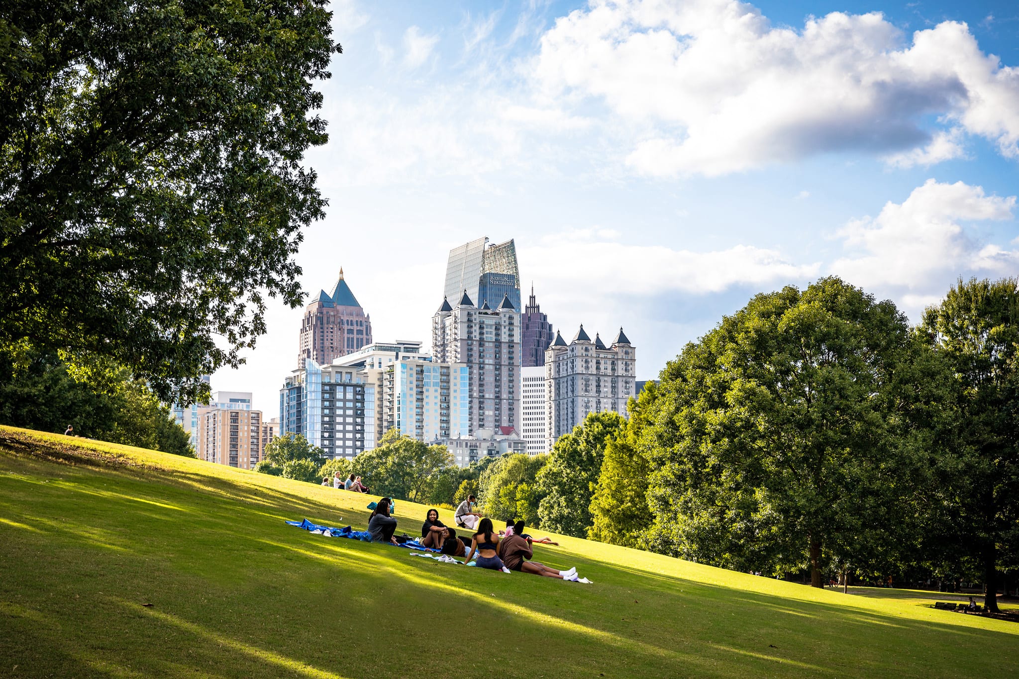 People relax on a grassy hill in a park, with a city skyline visible in the distance in Atlanta, Georgia.
