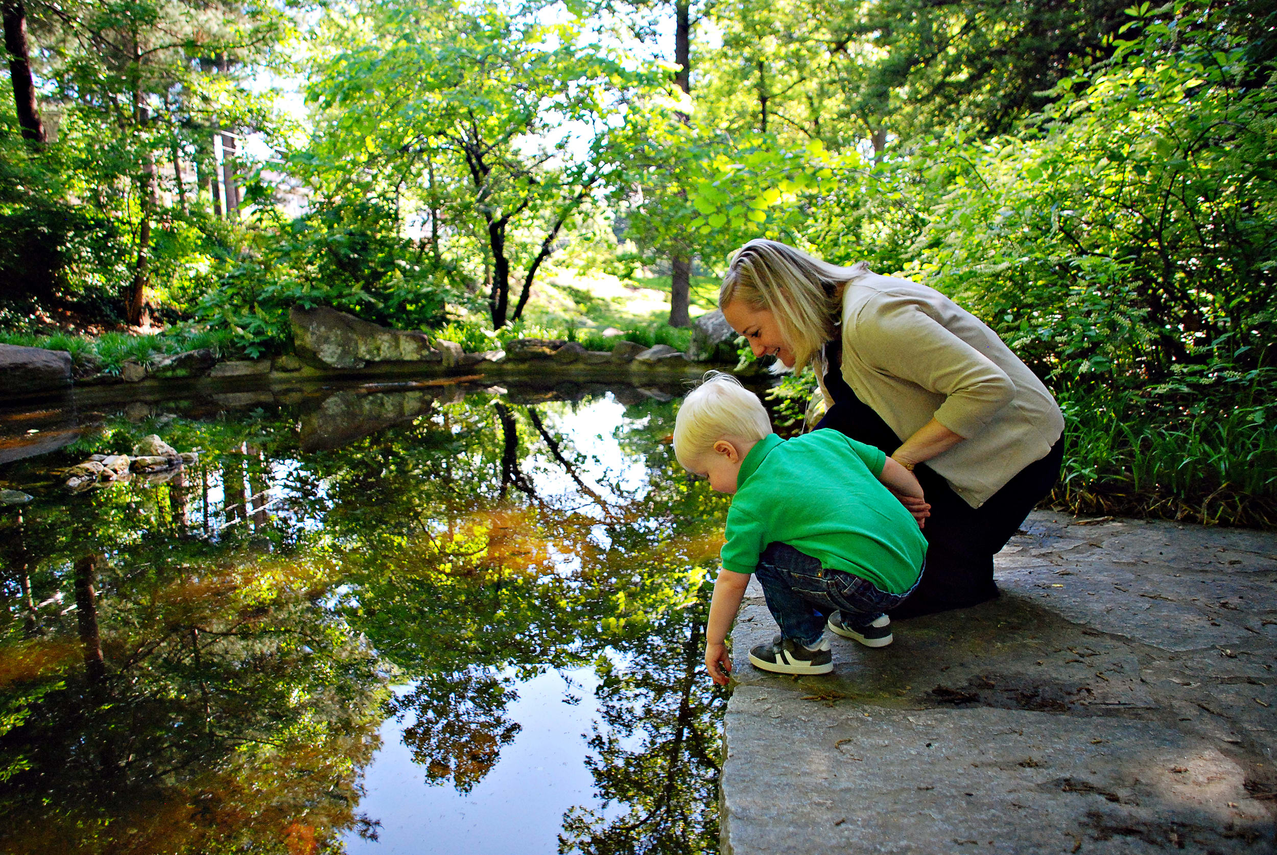 A woman and child kneel by a serene pond, reaching into the water amidst lush greenery in Atlanta, Georgia.