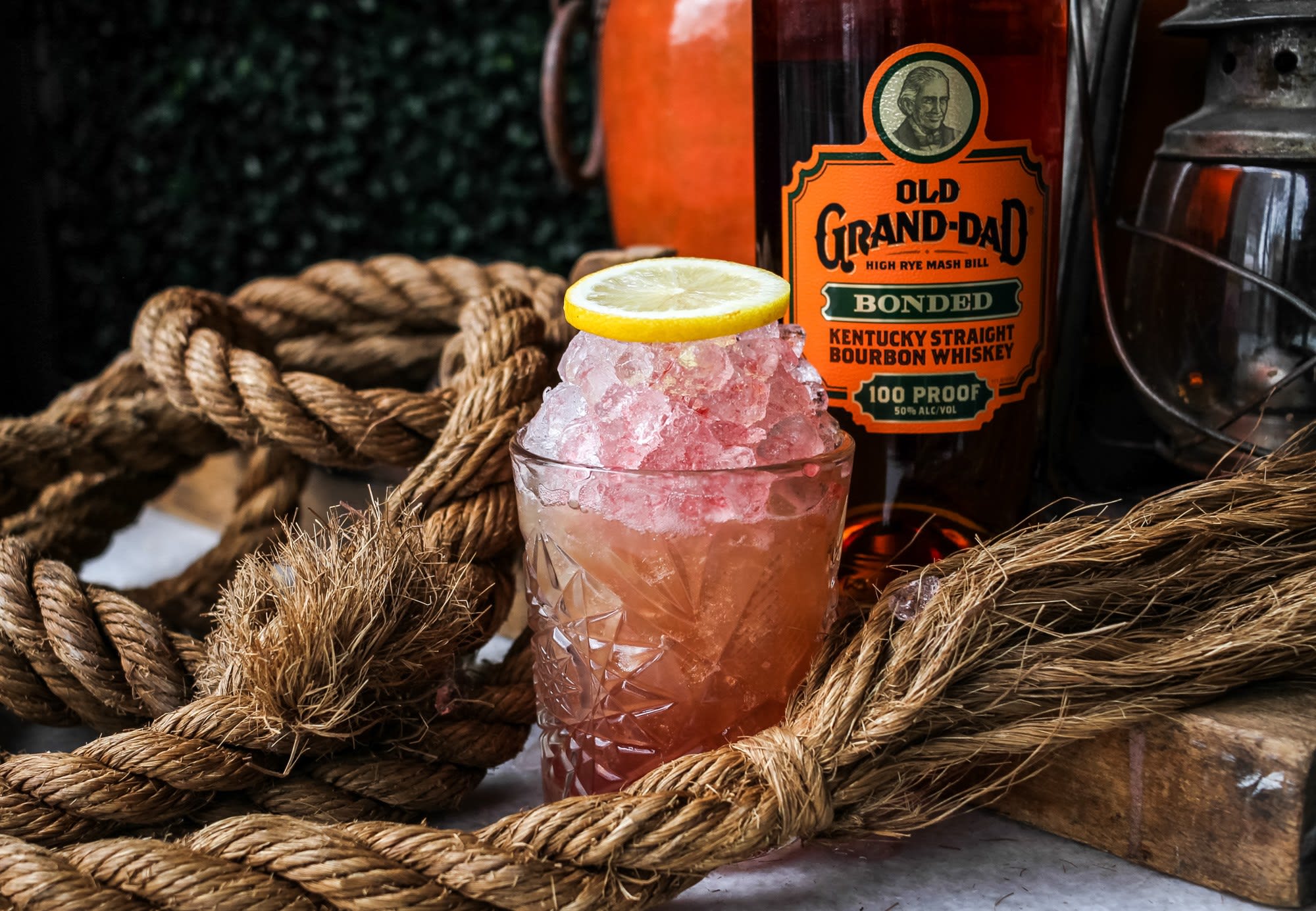 A glass filled with a pinkish-red cocktail, garnished with a lemon slice, sits on a table with a bottle of Old Grand-Dad Bonded Bourbon Whiskey and a pile of thick ropes in Atlanta, Georgia.