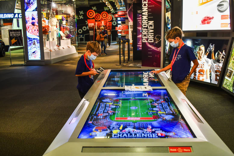Two boys wearing masks play an interactive football game on a large touchscreen display in Atlanta, Georgia.