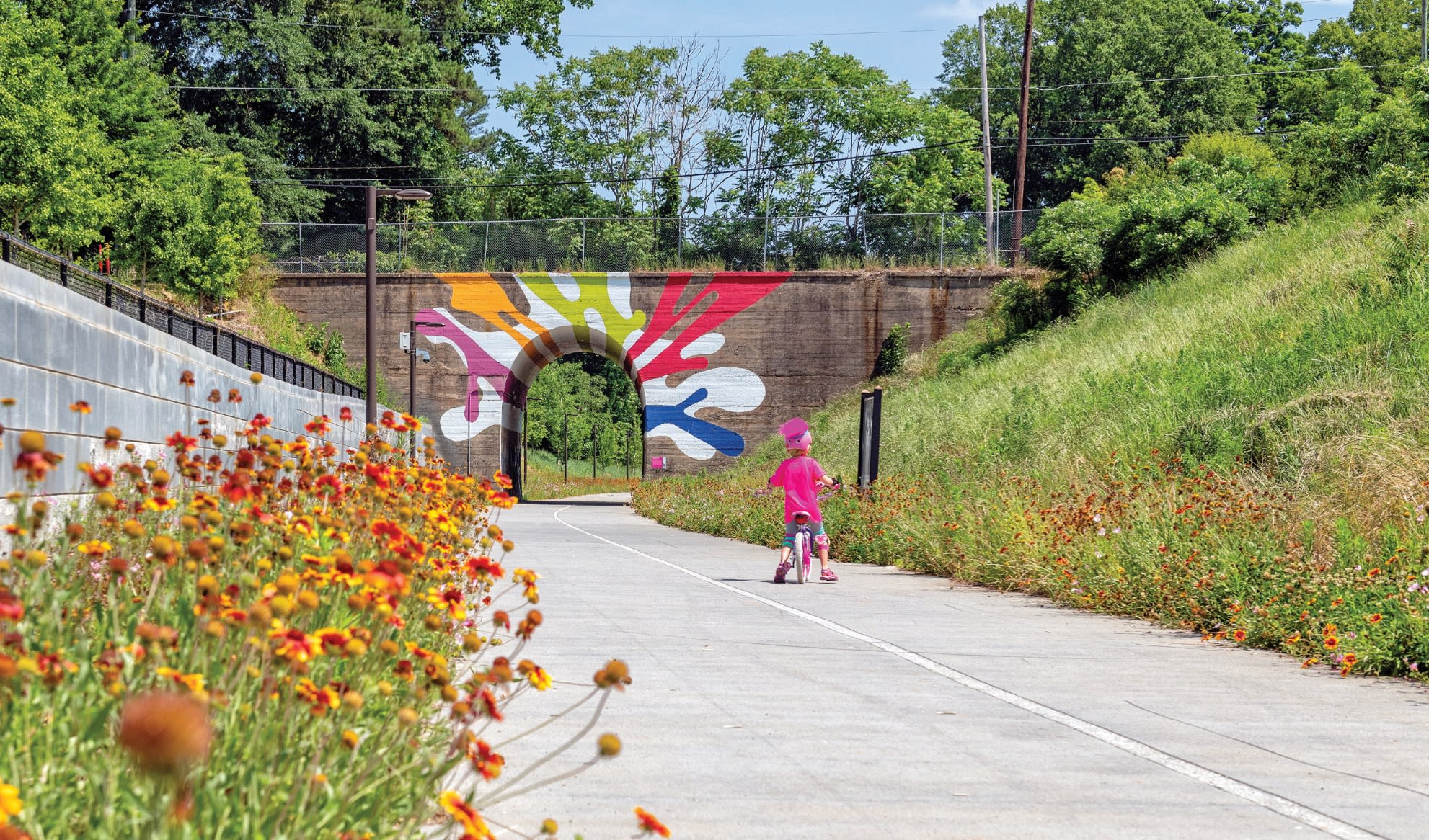 A child in a pink helmet rides a bike along a paved path surrounded by colorful flowers and a tunnel in Atlanta, Georgia.