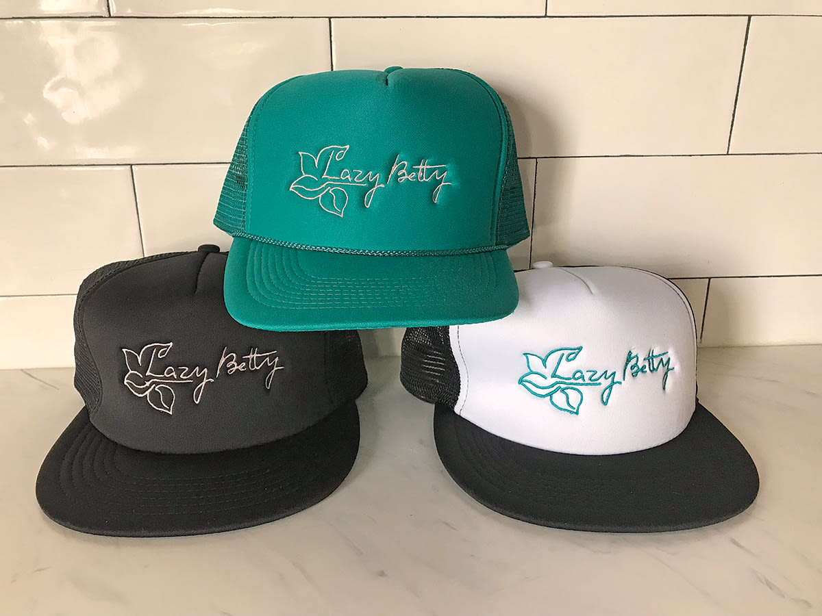 Three trucker hats in black, white, and teal with 'Lazy Betty' logo and leaf design in Atlanta, Georgia.