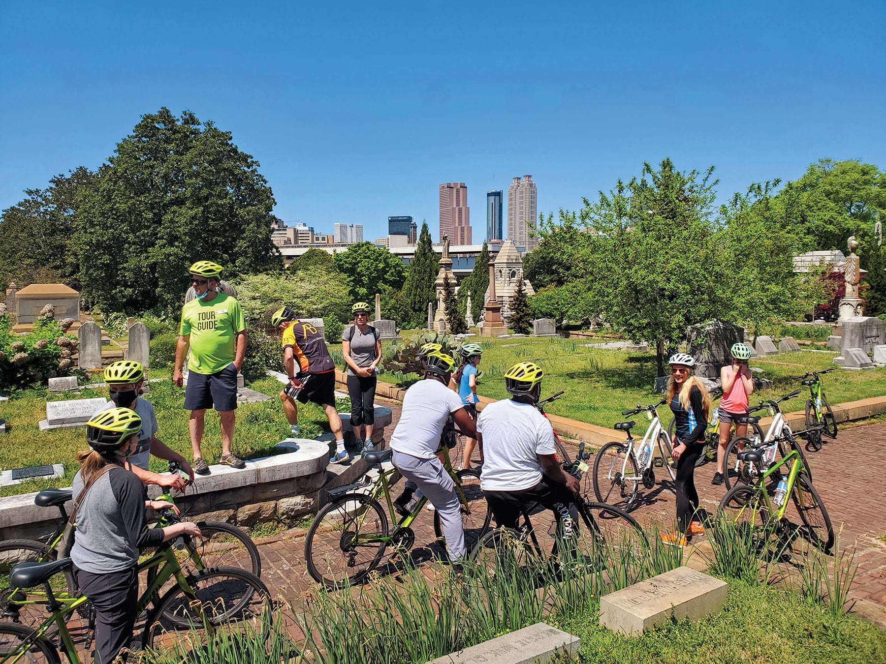 A group of cyclists wearing helmets and bright clothing ride through a cemetery with gravestones and trees in Atlanta, Georgia.
