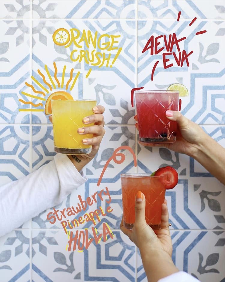 Three hands holding colorful cocktails with fruit garnishes against a patterned tile wall in Atlanta, Georgia.