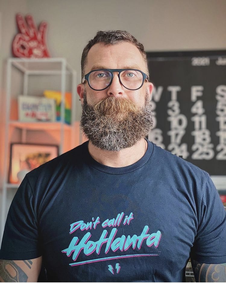 A bearded man with glasses wears a black t-shirt that says 'Don't Call It Hotlanta' in neon colors in Atlanta, Georgia.