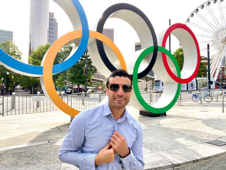 A man in sunglasses stands in front of the Olympic rings sculpture in a city park in Atlanta, Georgia.