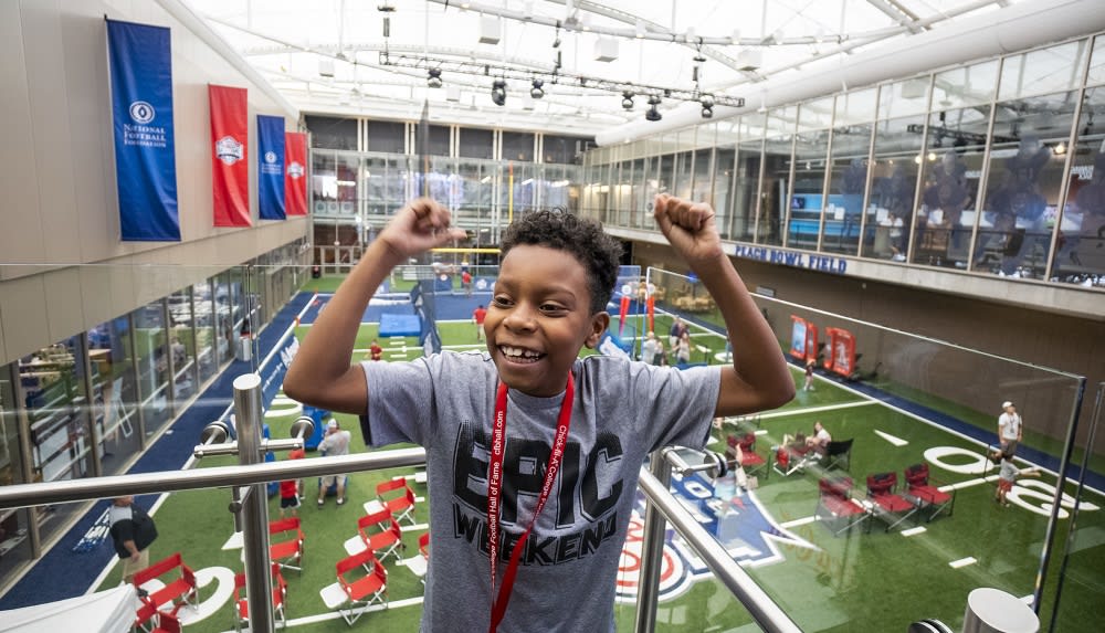 A young boy with a big smile stands on a balcony overlooking an indoor sports facility, celebrating with his arms raised in Atlanta, Georgia.