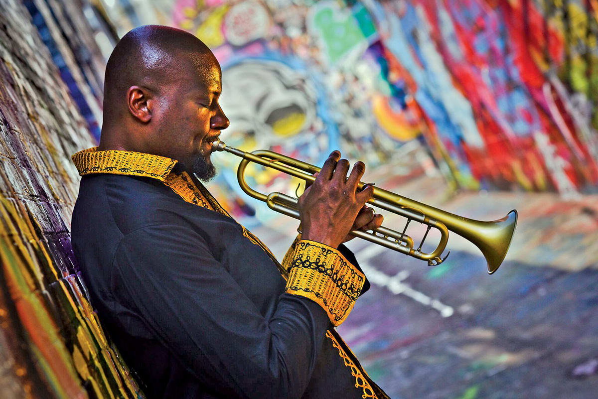 A bald man in a black and gold jacket plays a trumpet against a colorful graffiti wall in Atlanta, Georgia.