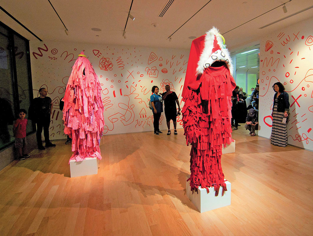 Two red, fringed costumes stand on pedestals in a brightly lit art gallery with colorful abstract wall murals in Atlanta, Georgia.