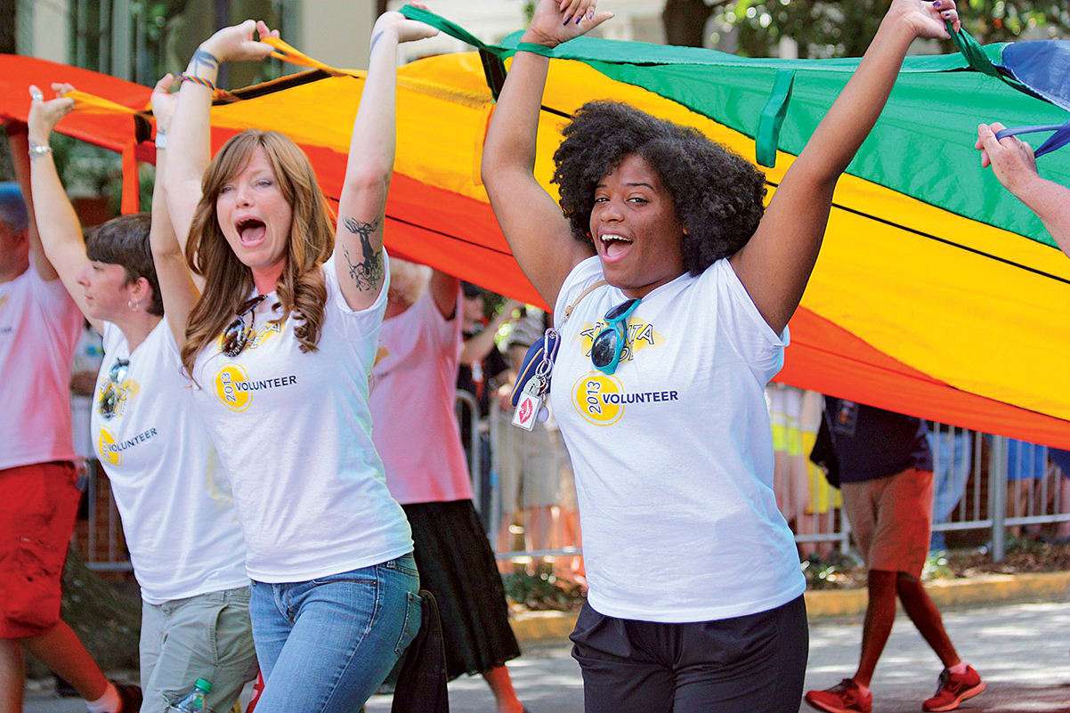 A group of enthusiastic volunteers joyfully hold up a large rainbow flag at a pride parade in Atlanta, Georgia.