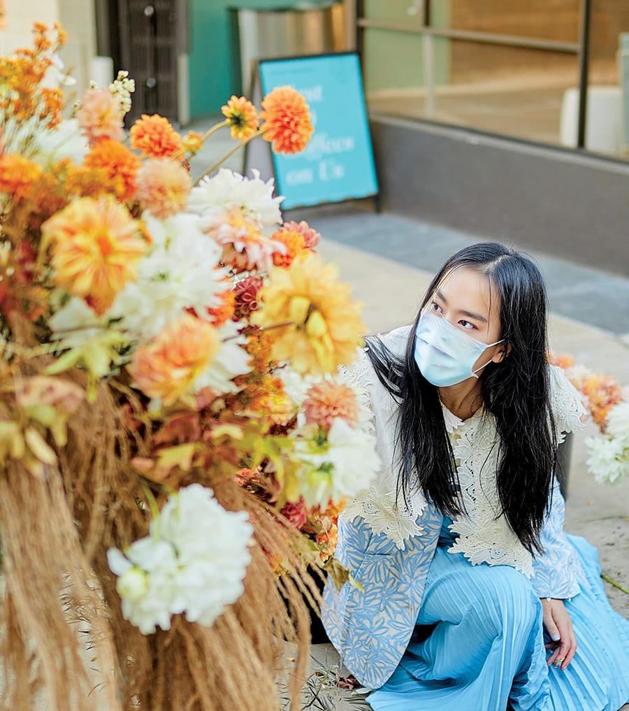 A young woman in a blue dress and mask kneels beside a vibrant bouquet of colorful flowers in Atlanta, Georgia.