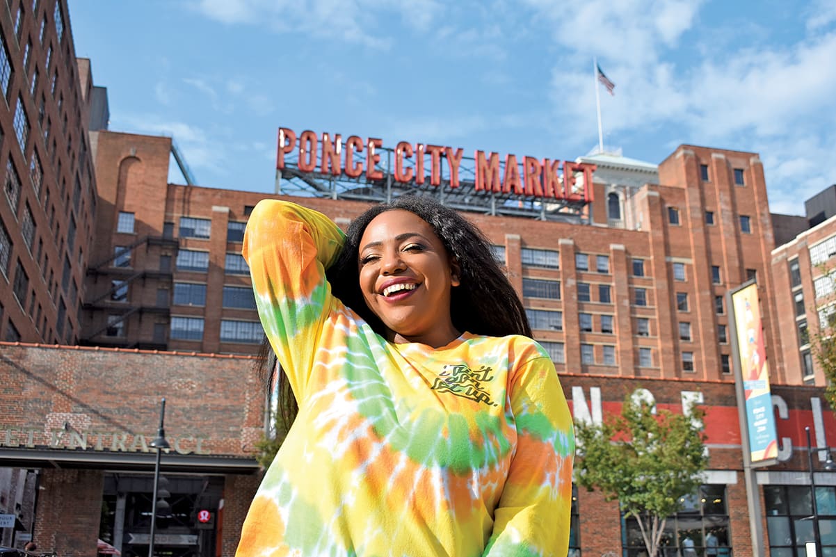 A woman in a colorful tie-dye shirt smiles in front of a large brick building with a sign in Atlanta, Georgia.