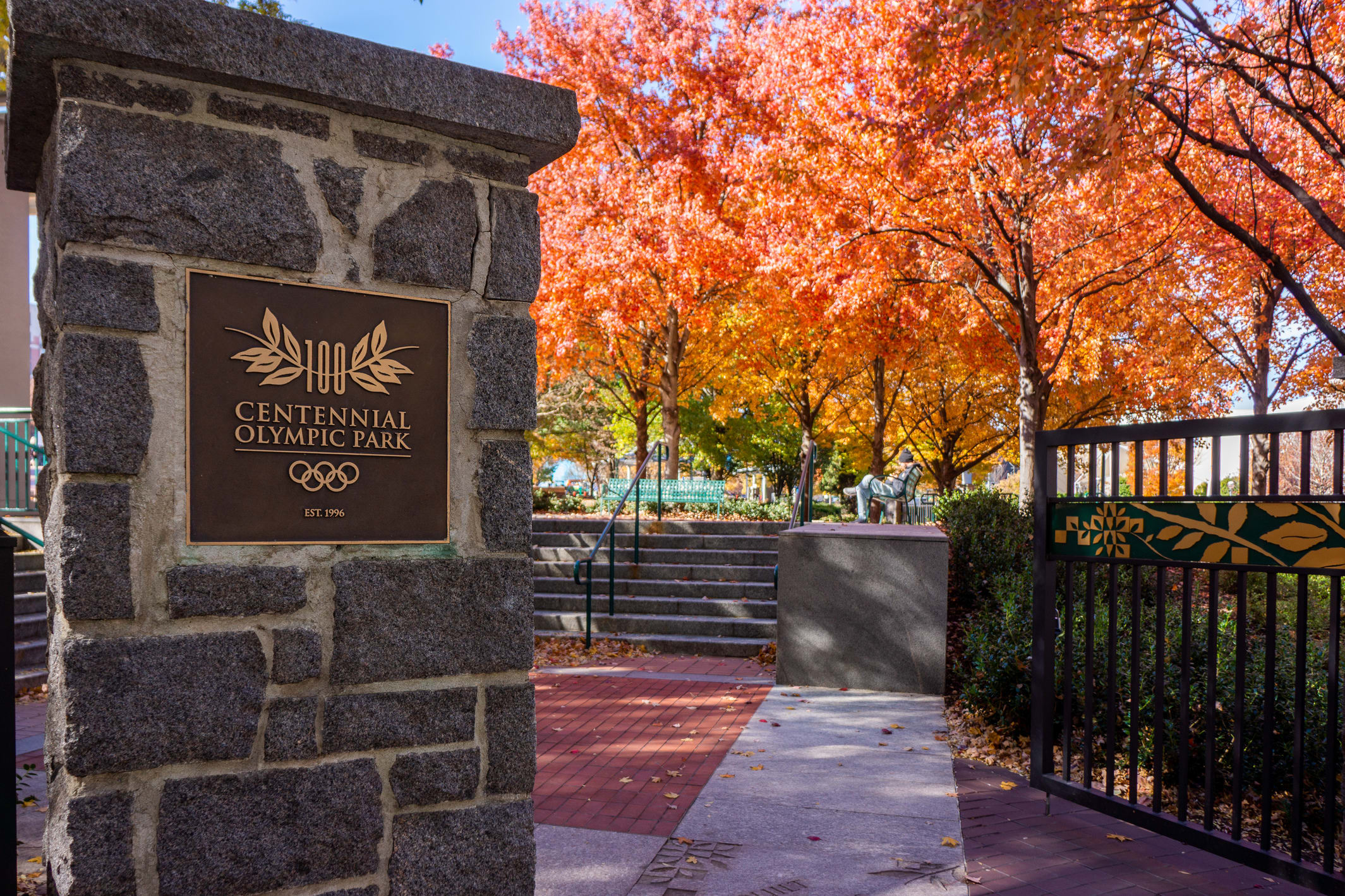 A stone pillar with a plaque marks the entrance to Centennial Olympic Park, surrounded by autumn foliage in Atlanta, Georgia.