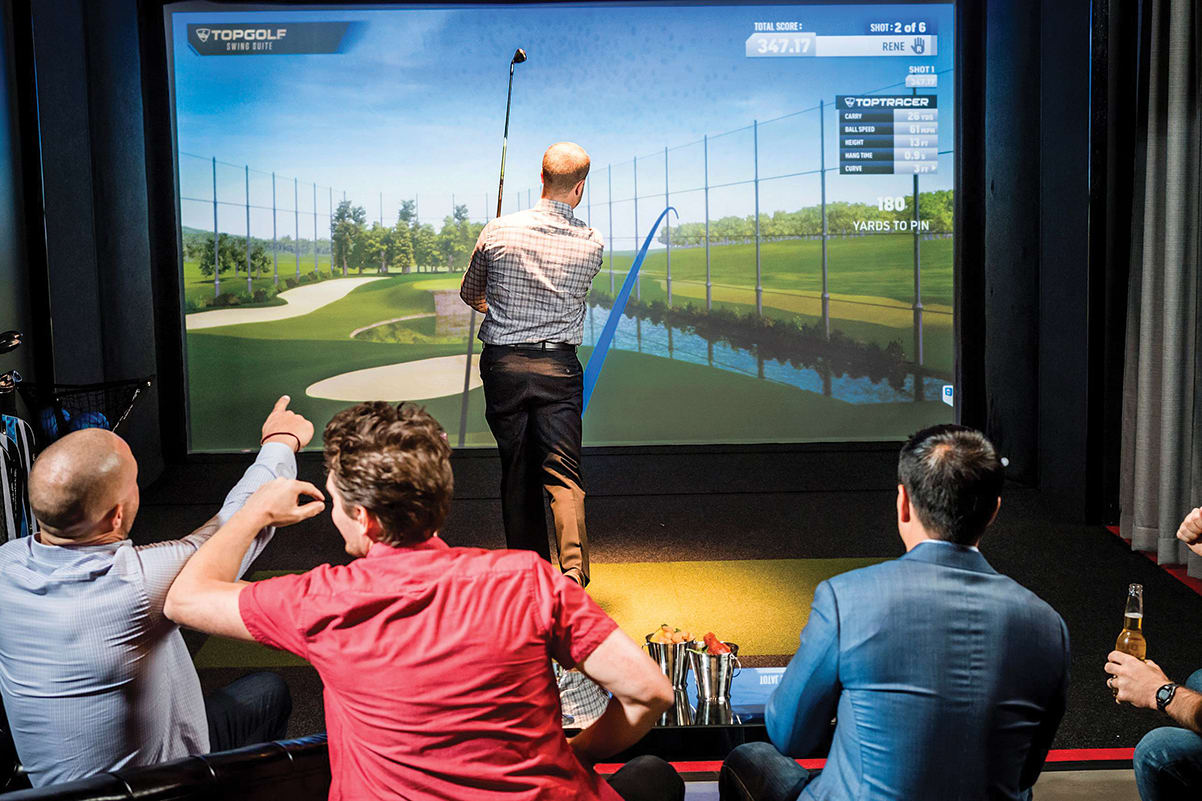 Men play virtual golf in a simulator room, watching a screen display a golf course in Atlanta, Georgia.