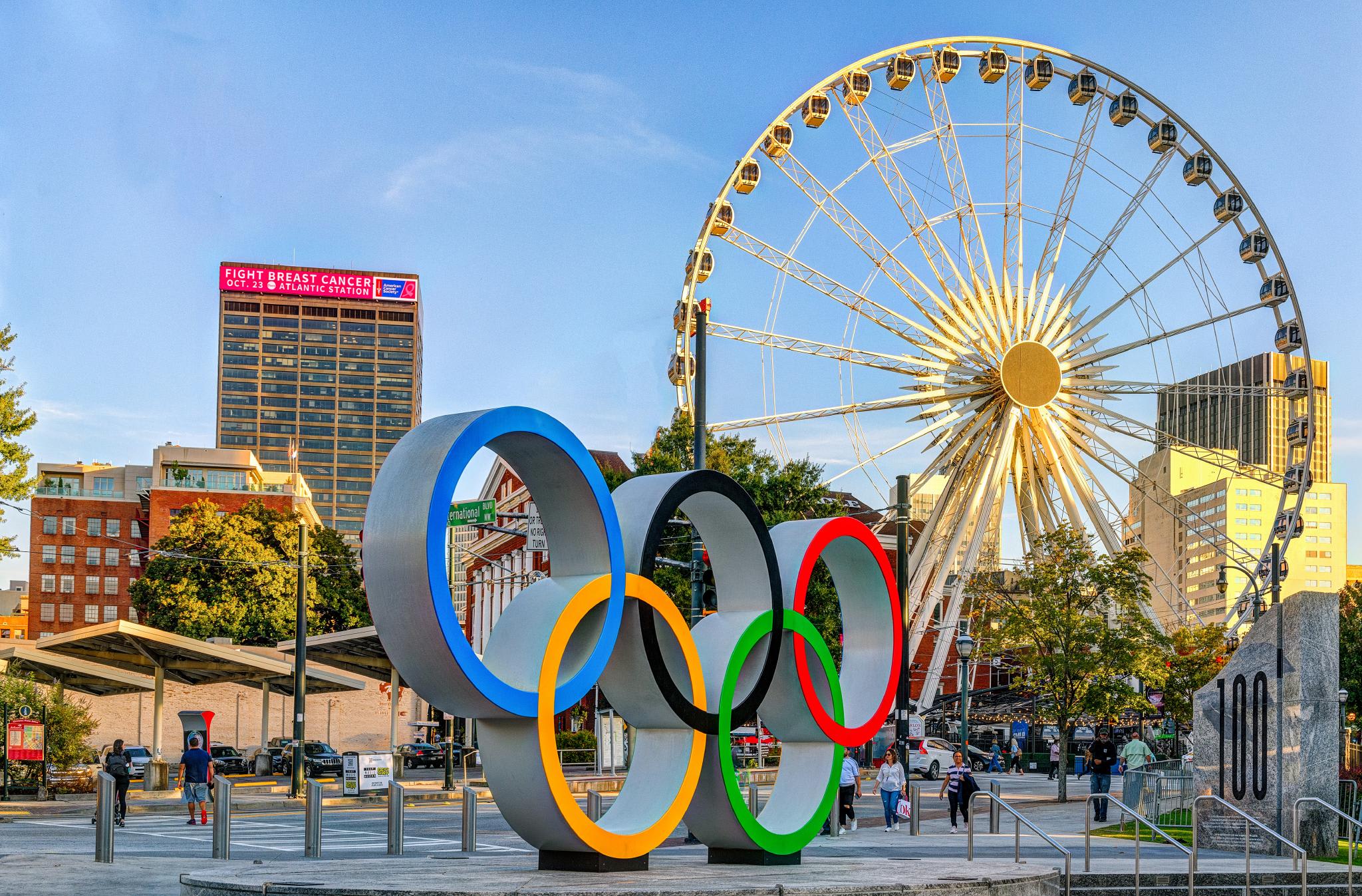 A cityscape with a large Ferris wheel, Olympic rings, and a tall building with a pink sign in Atlanta, Georgia.