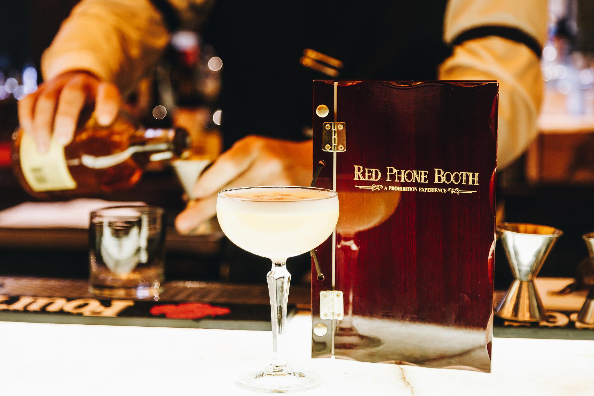 A bartender pours a drink into a martini glass at a bar with a 'Red Phone Booth' menu in Atlanta, Georgia.