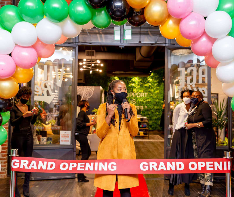 A woman in a yellow coat stands on a red carpet, holding a microphone at a grand opening event in Atlanta, Georgia.