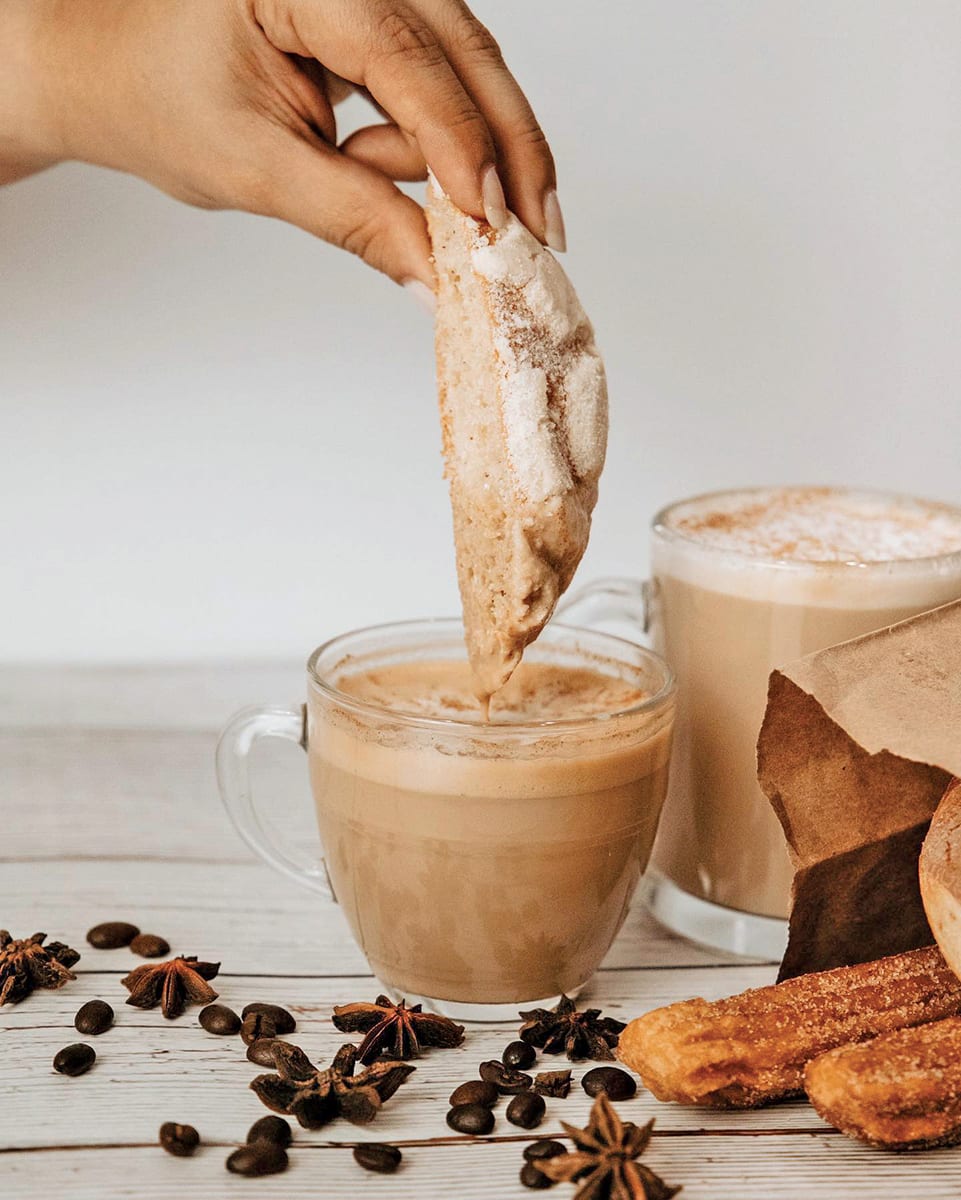 A hand dips a sugar-coated cookie into a frothy latte, surrounded by coffee beans and pastries in Atlanta, Georgia.