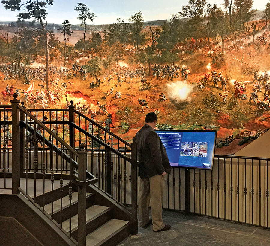A man stands before a large mural depicting a Civil War battle scene with soldiers and cannons in Atlanta, Georgia.