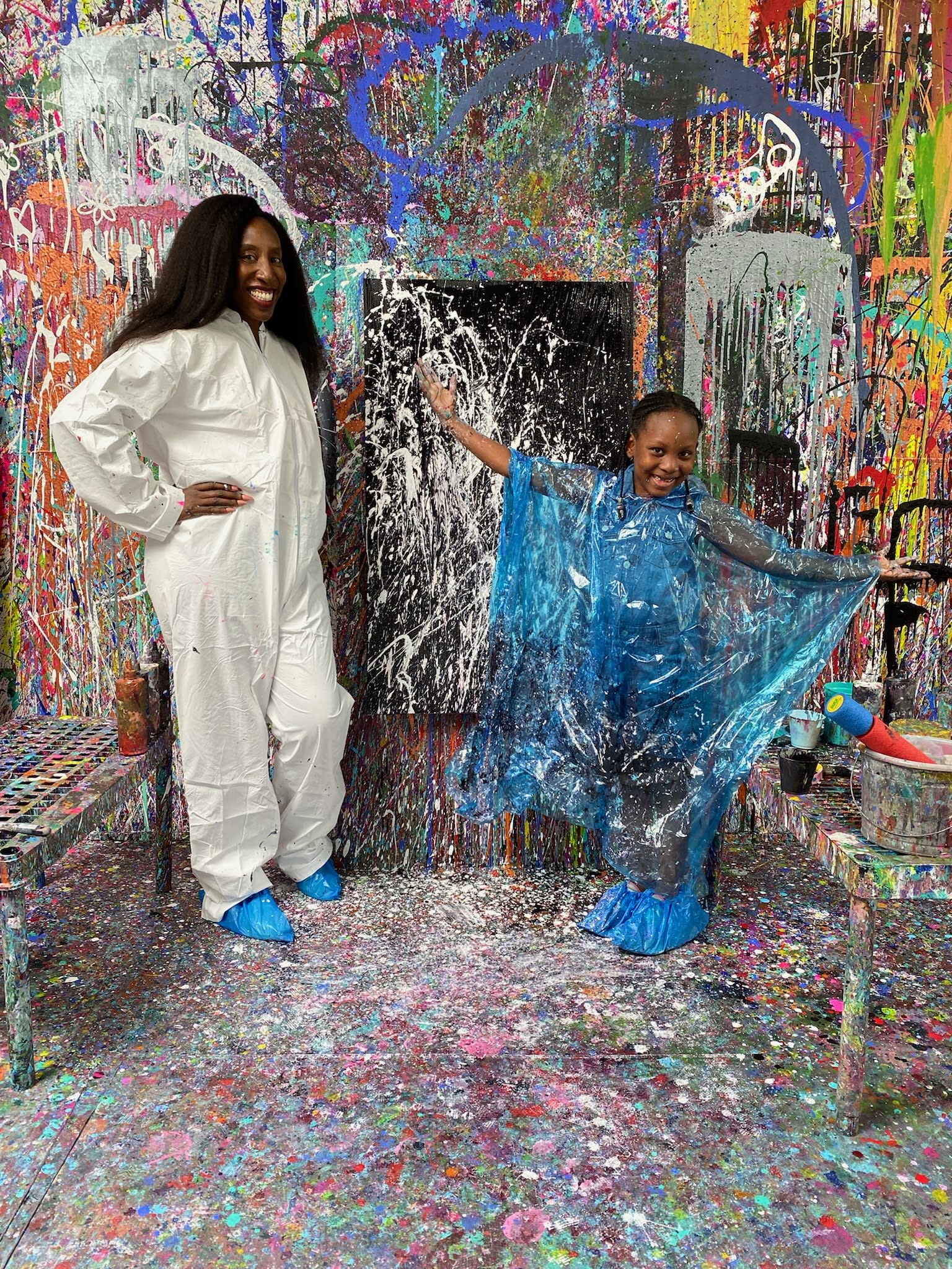 Two women in white coveralls and blue boots stand amidst a colorful paint-splattered room in Atlanta, Georgia.