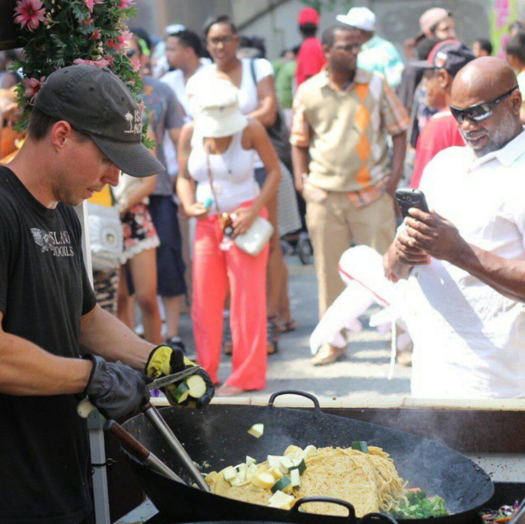 A chef in a black cap and gloves stirs vegetables in a large wok at an outdoor food event in Atlanta, Georgia.